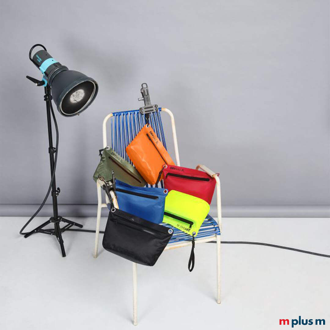 Verschiedene Drybags (wasserdichte Taschen) von Halfar in den Farben Schwarz, Orange, Rot, Gelb-Grün, Blau und Olivgrün, liegen auf einem Vintage-Stuhl in einem Fotostudio-Setting mit Beleuchtung.